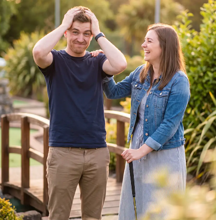 A couple on a date in the evening sun: he holds his head in both hands after a missed putt while she laughs with a hand on his shoulder, a little wooden bridge behind them