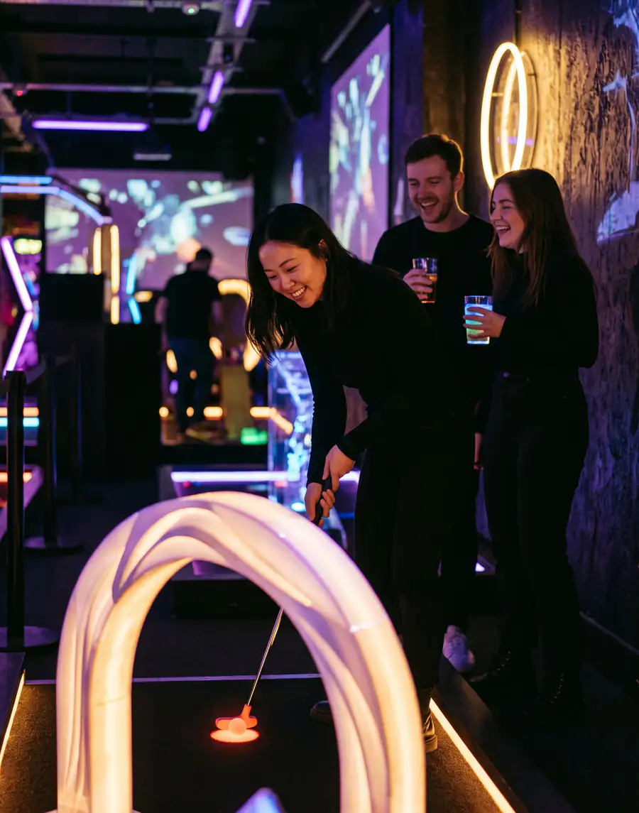 A woman putts through a glowing arch on an indoor neon course while two friends with drinks laugh behind her