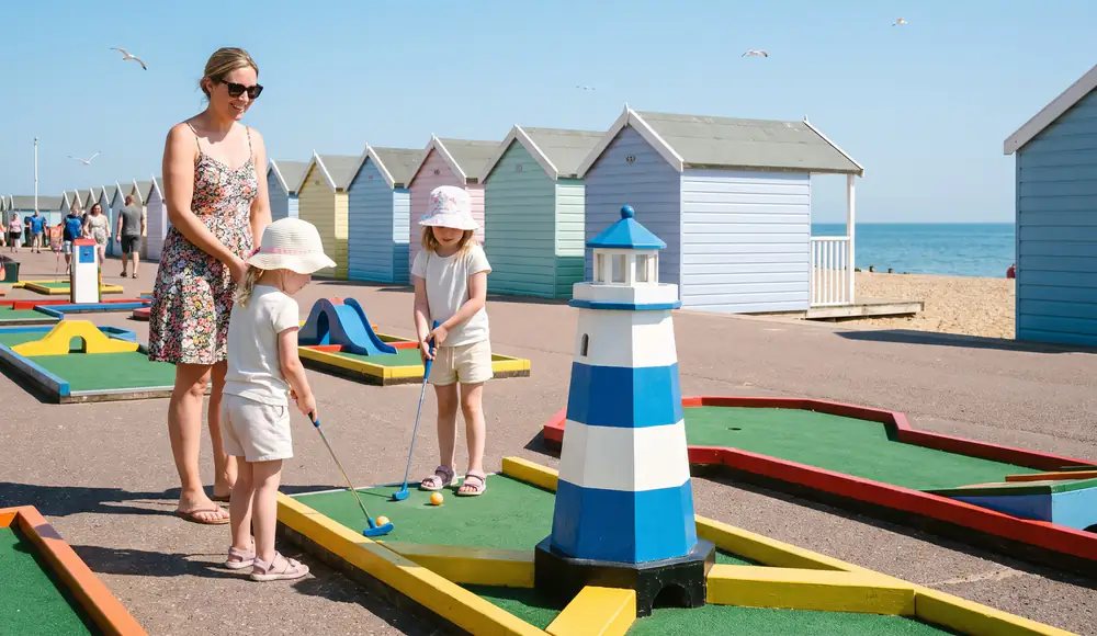 Two little girls in sun hats putt beside a blue and white lighthouse on a seafront course, with pastel beach huts and the sea behind