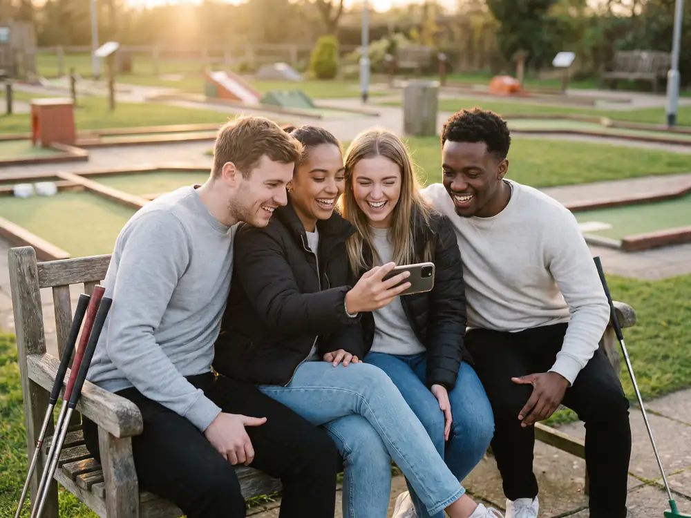 Four friends squeezed together on a bench beside the course at sunset, laughing at the result on one phone, their putters leaning on the bench