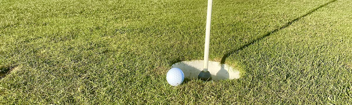 Close up of a wide cup cut into the grass, with the flagstick standing in it and a golf ball resting on the lip