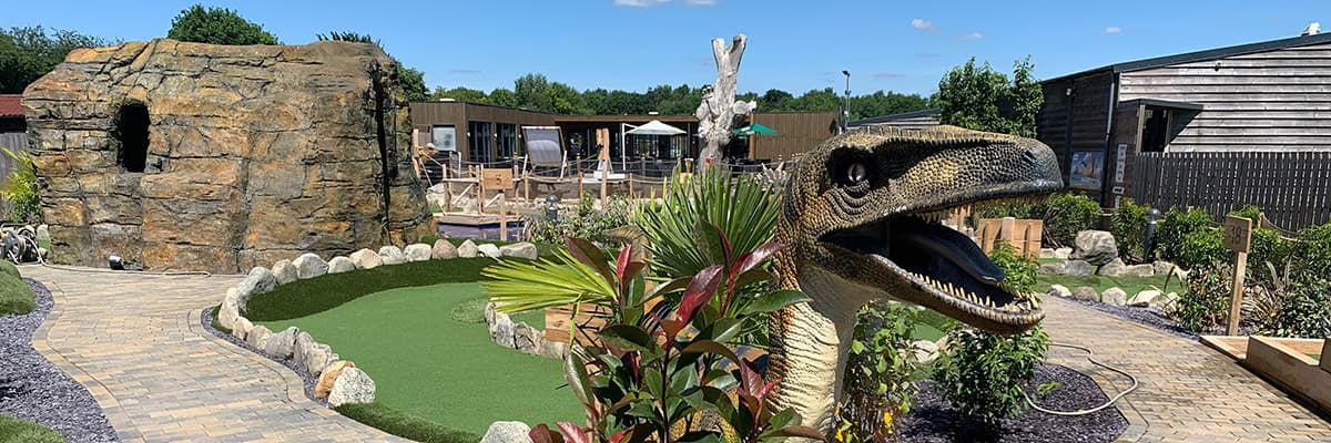 The dinosaur course on a bright day: a tall rock face with a cave mouth, a green ringed with boulders, a life-sized raptor and the cafe decking behind